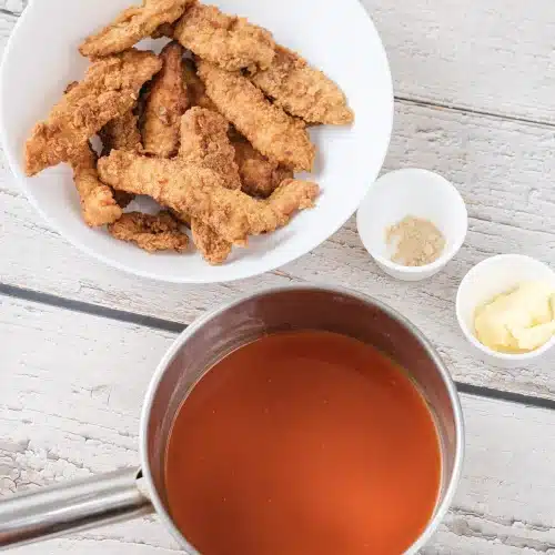 Baked breaded strips in a bowl beside a saucepan of orange buffalo sauce, butter and garlic powder