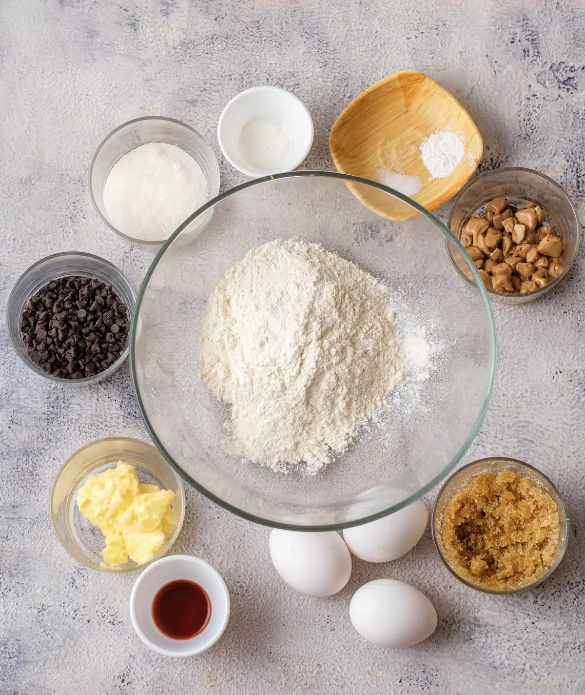 Flour in a glass bowl surrounded by butter, brown sugar, white sugar, eggs, vanilla, baking soda, salt and two bowls of baking morsels