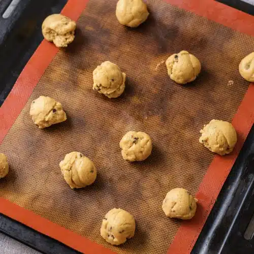 Scoops of cookie dough spaced out on a silicone mat on a dark baking sheet