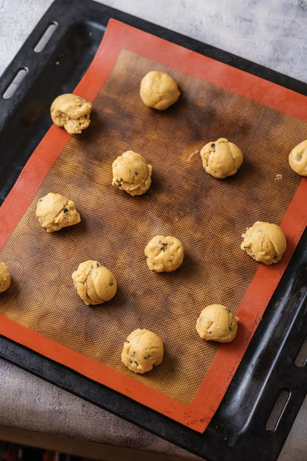 Balls of butterscotch chocolate chip cookie dough spaced out on a silicone mat on a dark baking sheet