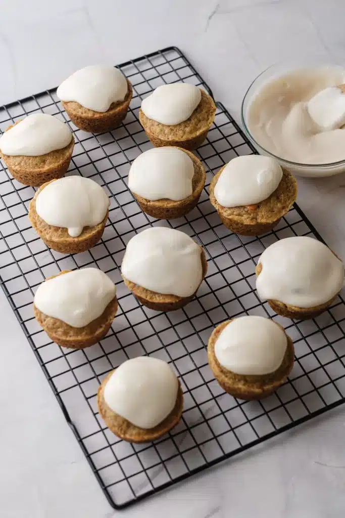 Carrot cake cupcakes with cream cheese frosting on a wire rack, seen from directly above with a bowl of frosting