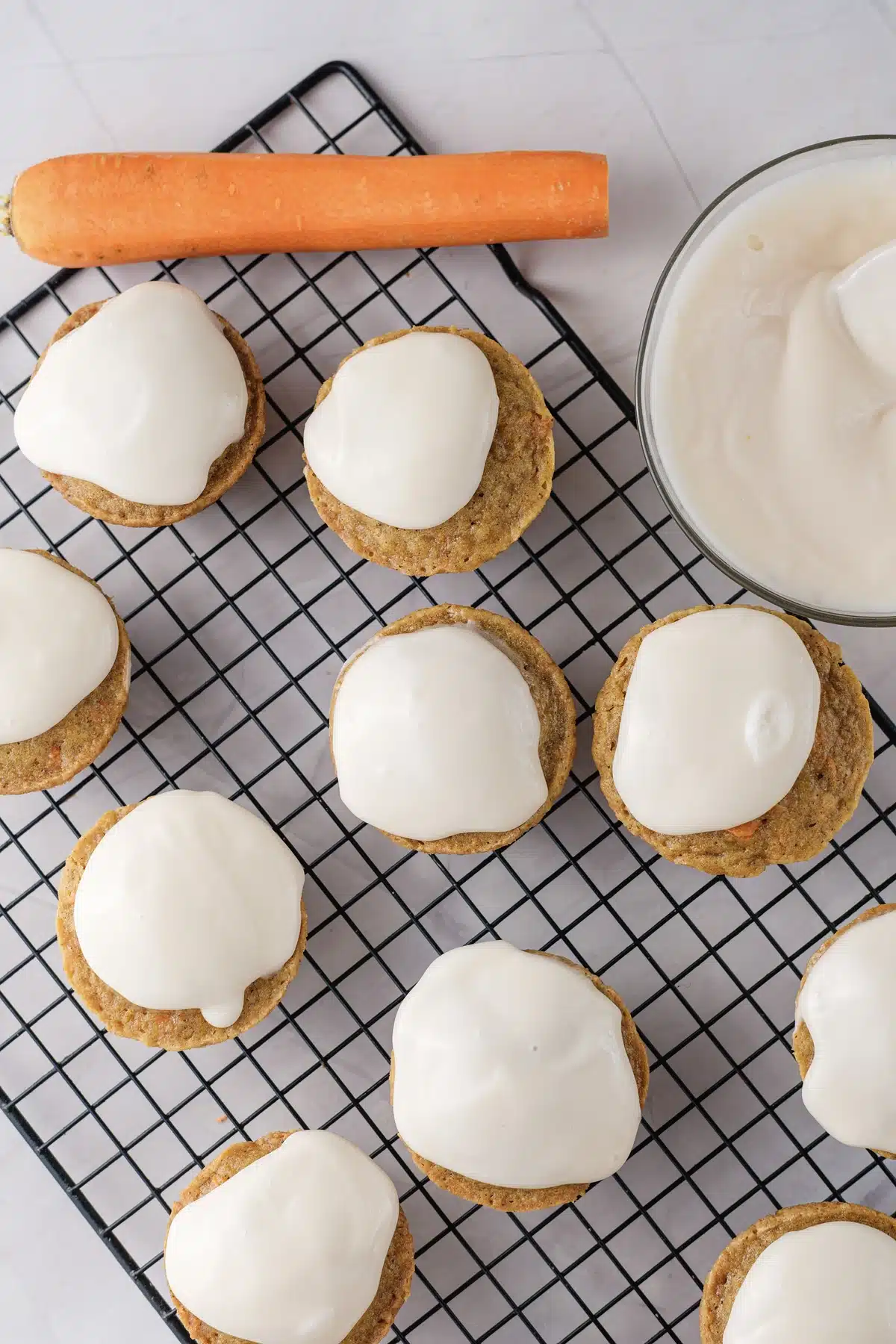 A dozen frosted rounds on a black wire rack from above with a whole carrot laid along the top
