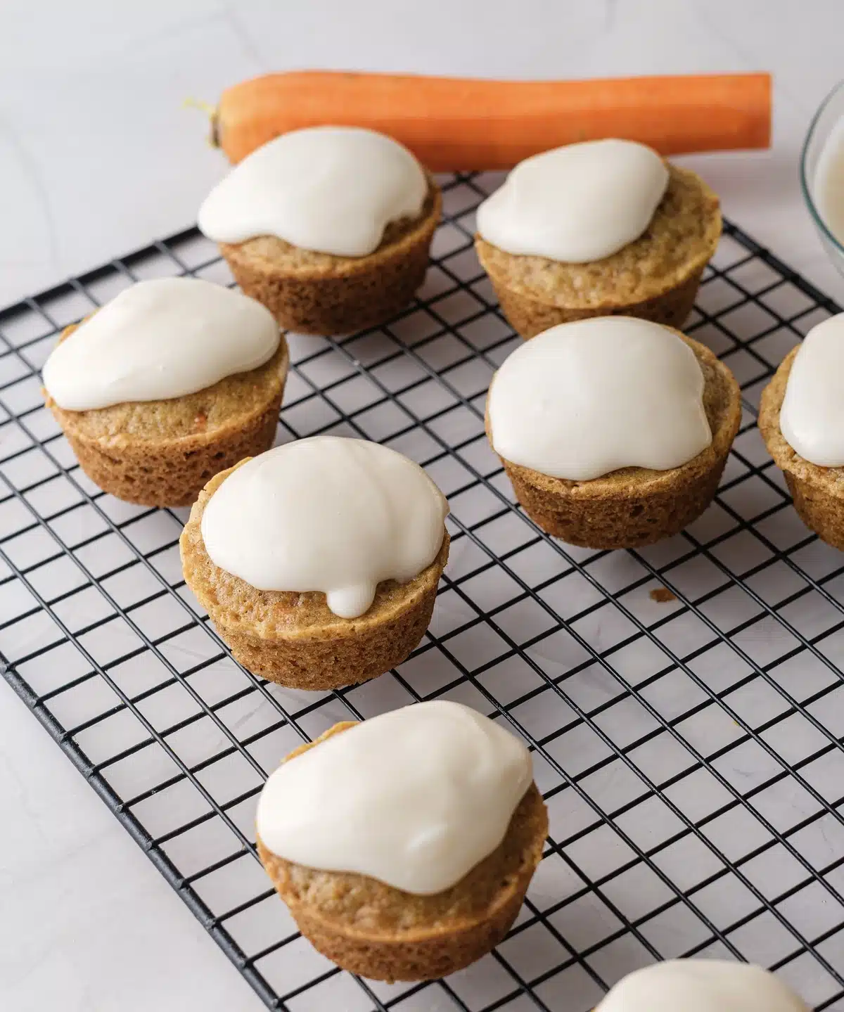 Carrot cake cupcakes with cream cheese frosting on a wire rack beside a whole carrot