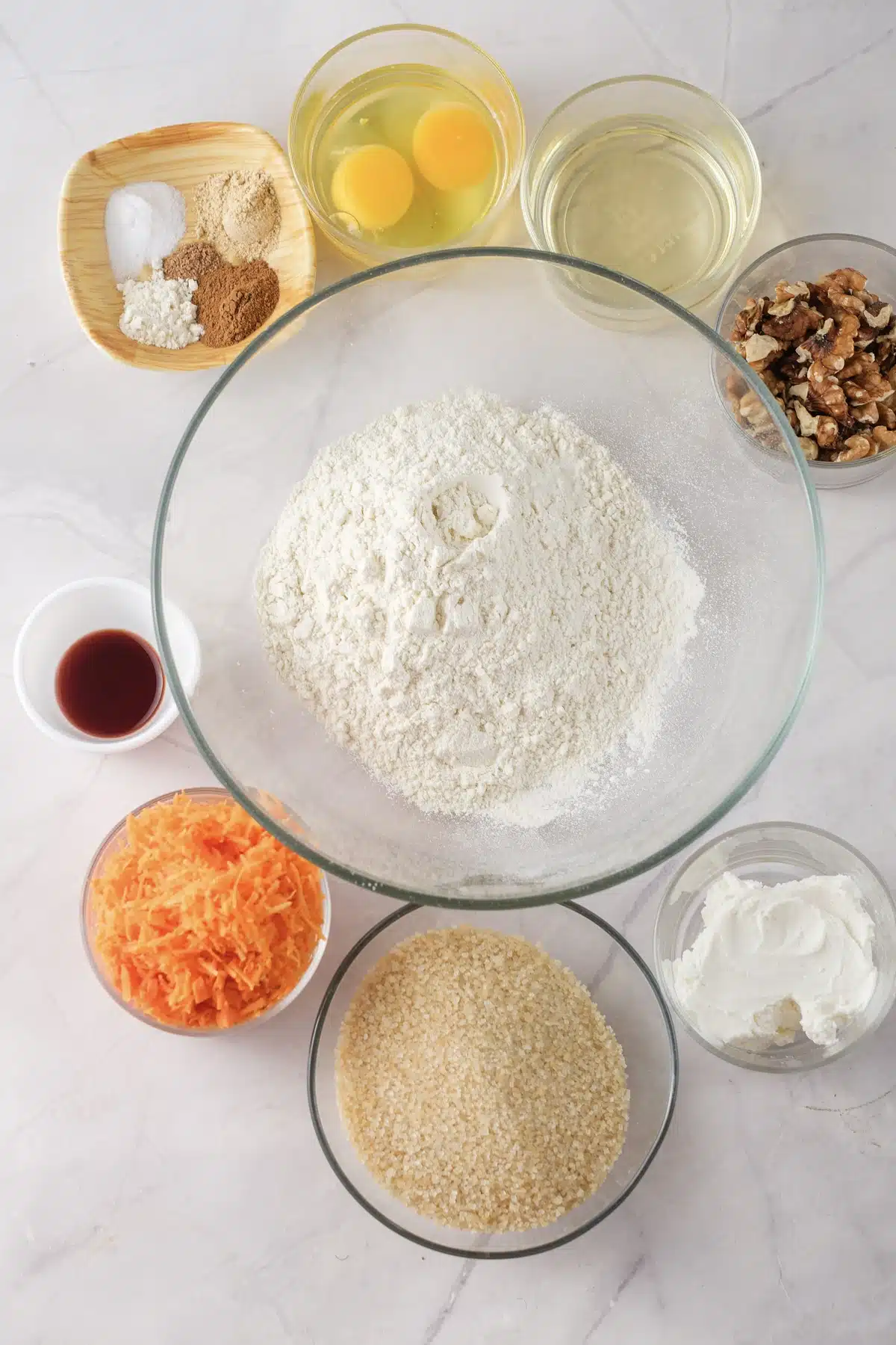 Flour in a glass bowl surrounded by grated orange shreds, brown sugar, eggs, oil, spices, vanilla, chopped walnuts and a dish of white frosting
