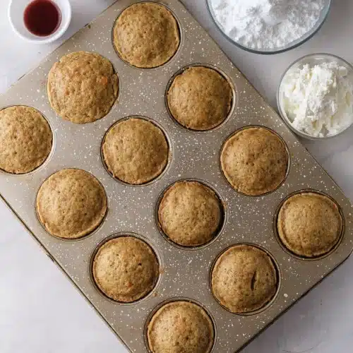 Twelve golden domed bakes in a bare muffin tin with powdered sugar and frosting beside it