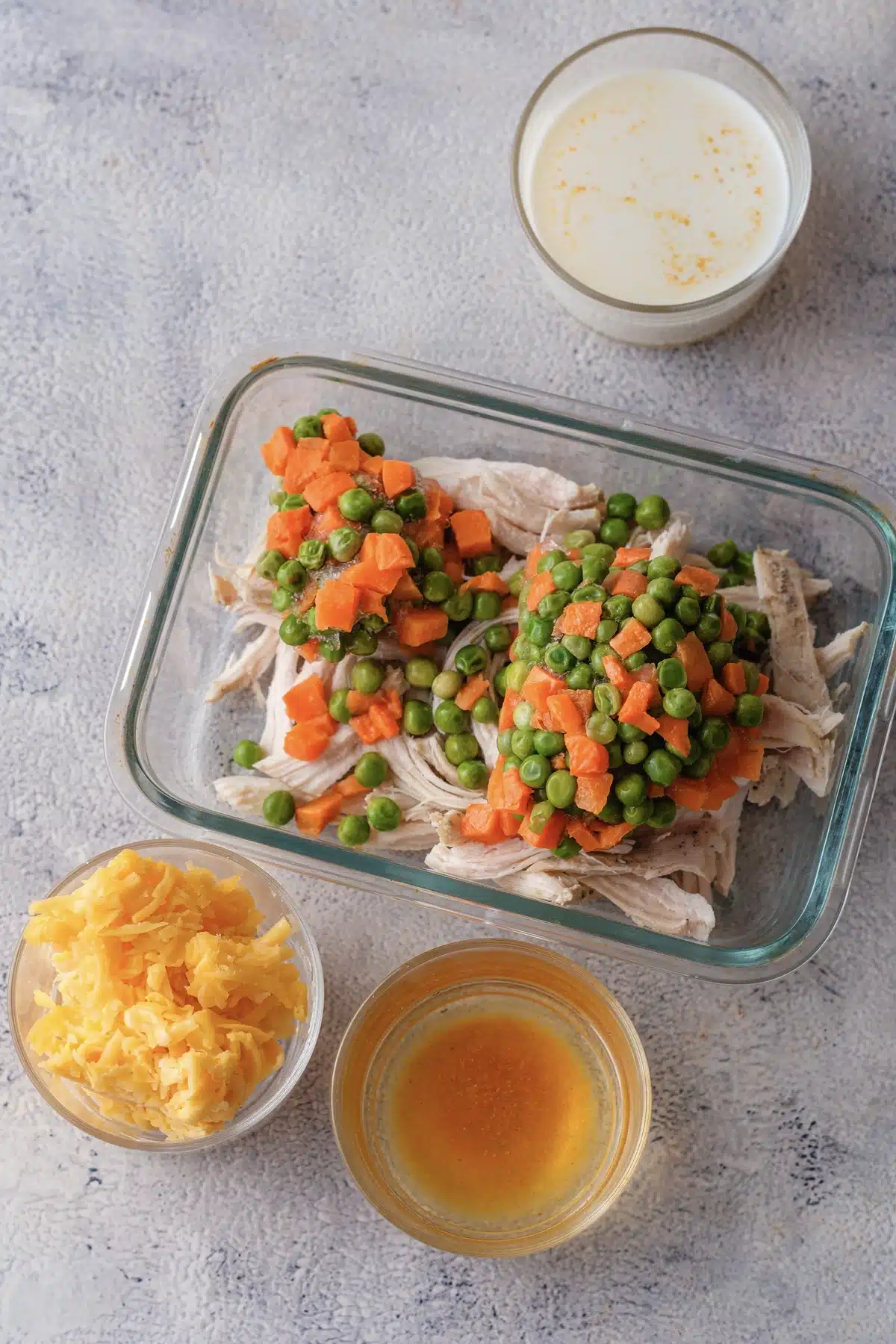 Peas and carrots piled over pulled meat in a glass dish, with a bowl of grated cheese and a bowl of broth beside it