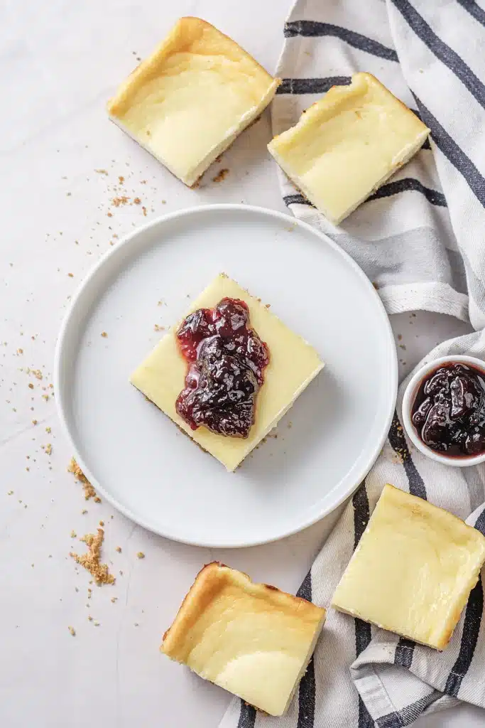 Cherry cheesecake bars plated on white with more squares on a linen cloth