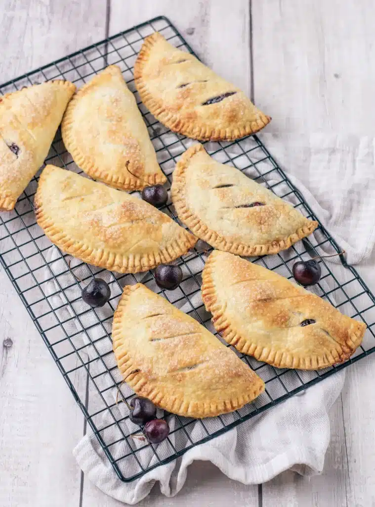 Cherry hand pies on a wire rack seen from directly above, sugar-topped with fresh cherries between them