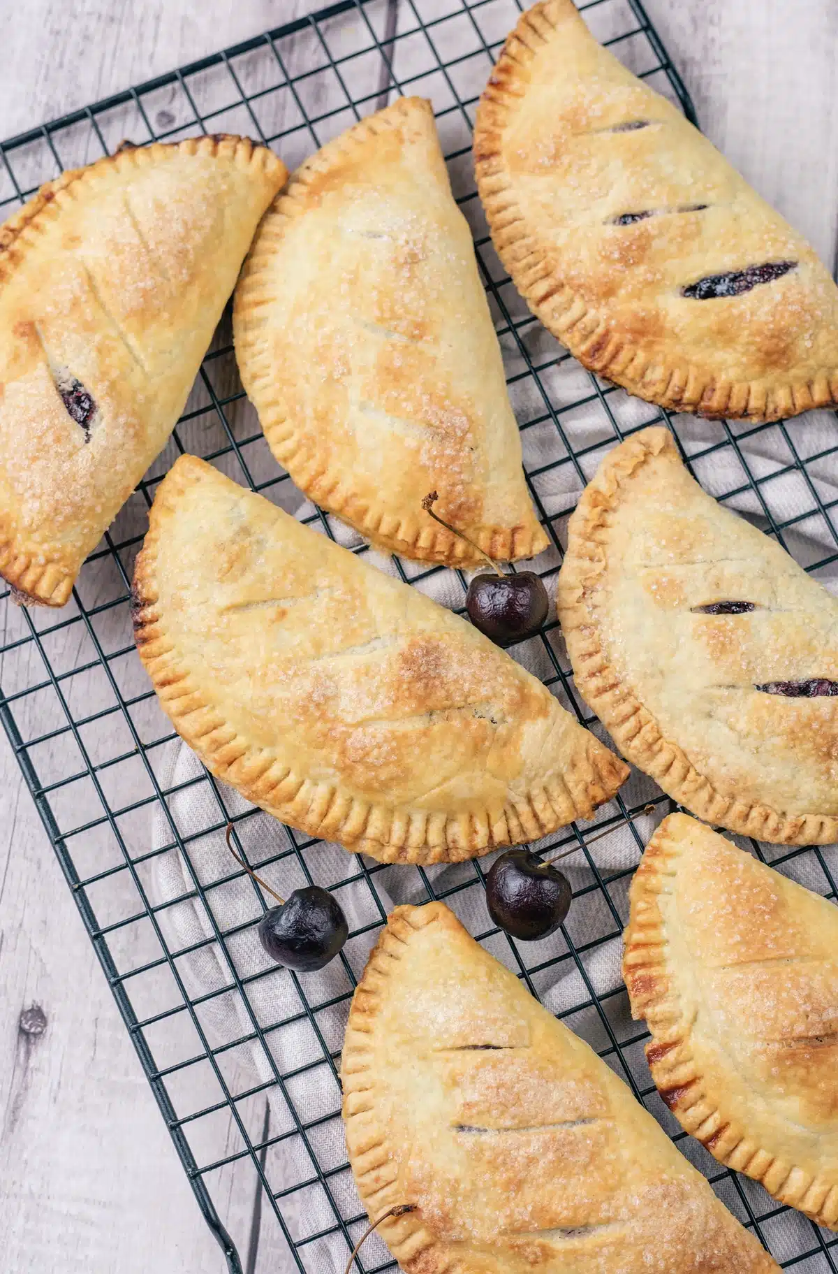 Sugar-topped half-moon pastries fanned across a wire rack with cherries and a striped cloth
