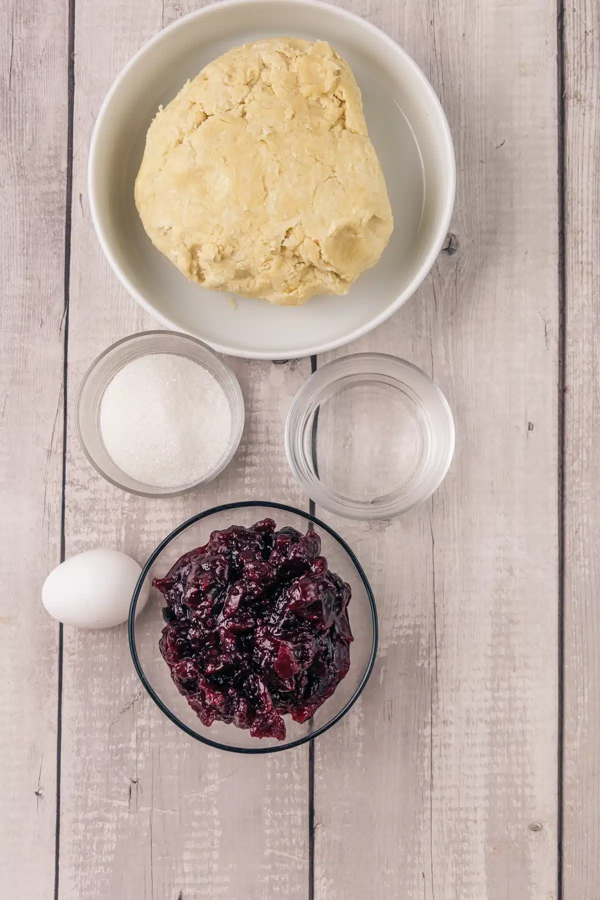 A ball of pale pastry dough in a bowl with coarse sugar, water, an egg and a bowl of dark fruit filling
