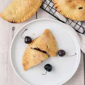 A single cherry hand pie split in two on a white plate with fresh cherries