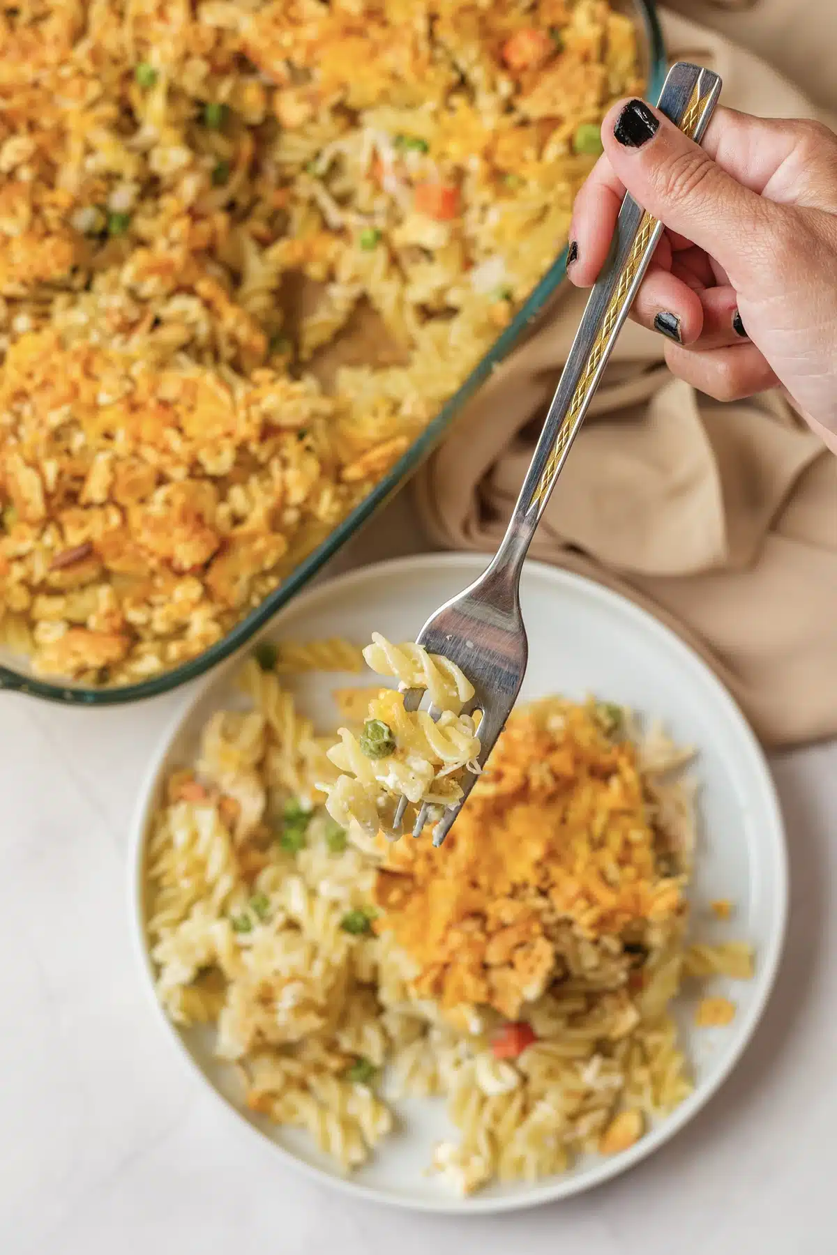 A fork lifting a bite of spiral pasta from a plated square, with the baking dish behind