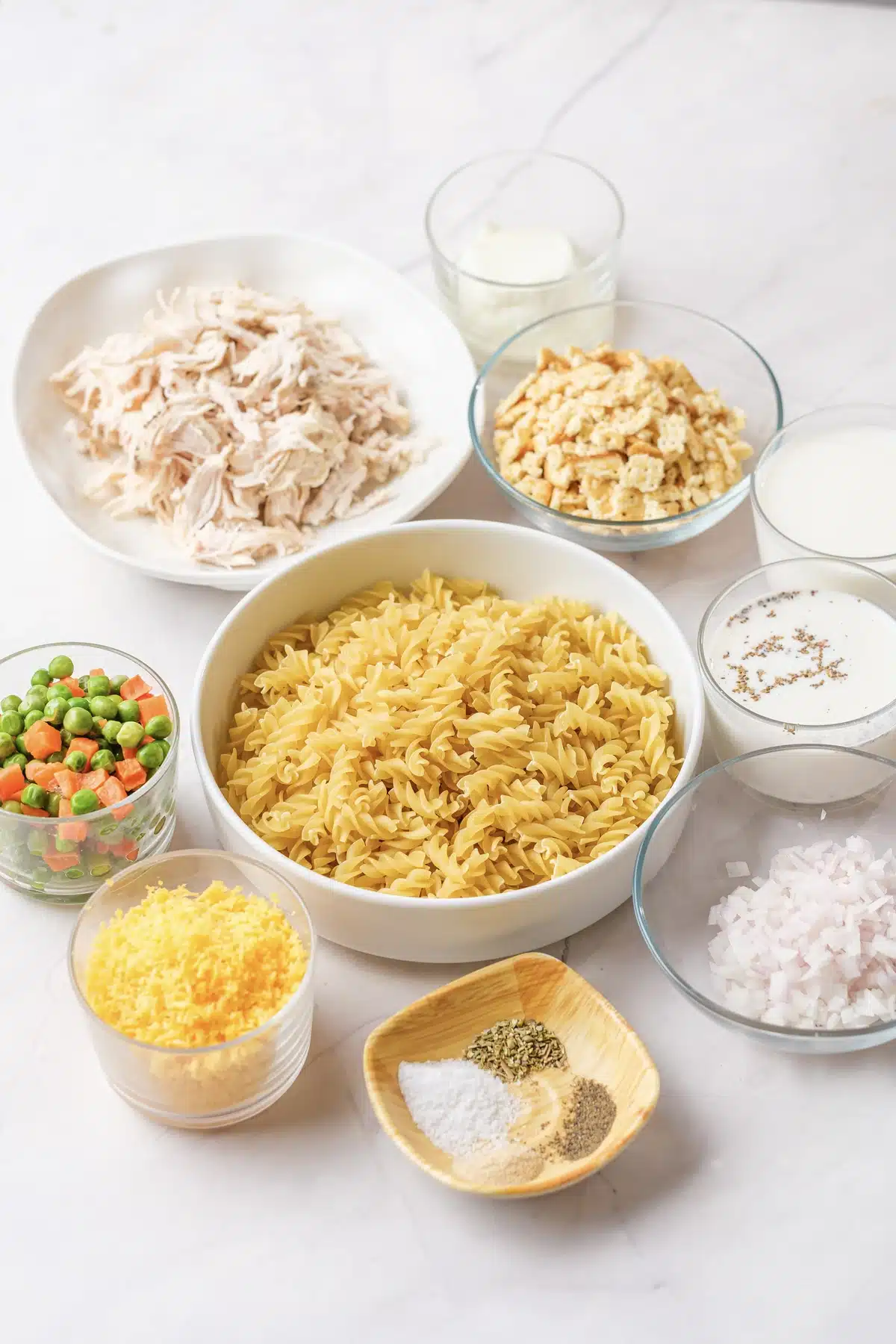 Shredded chicken, dry rotini, crushed crackers, sour cream, milk, cream of chicken soup, peas and carrots, cheddar, onion and seasonings in bowls