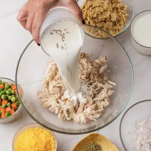 Cream of chicken soup poured from a measuring cup over shredded chicken in a glass bowl
