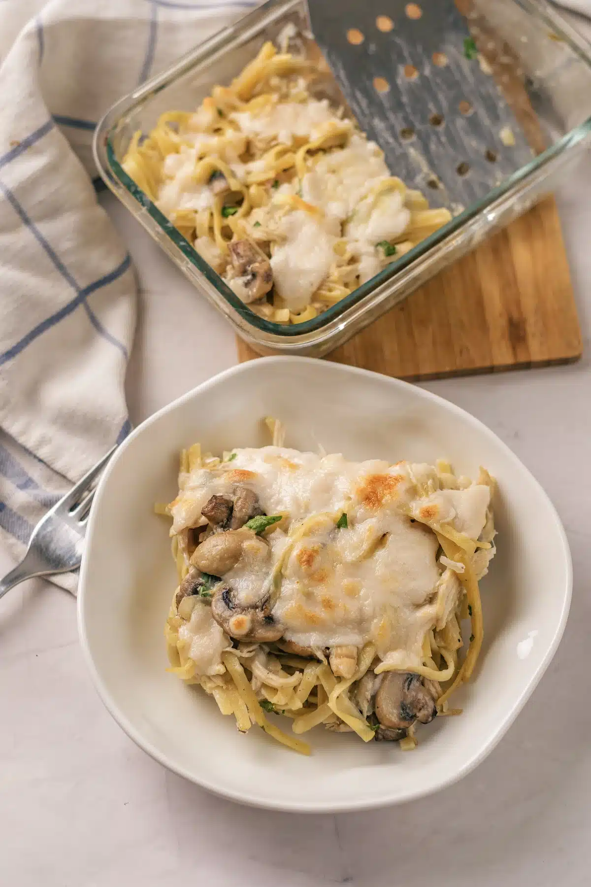 A plated square of chicken tetrazzini with the glass dish and a checked cloth behind it