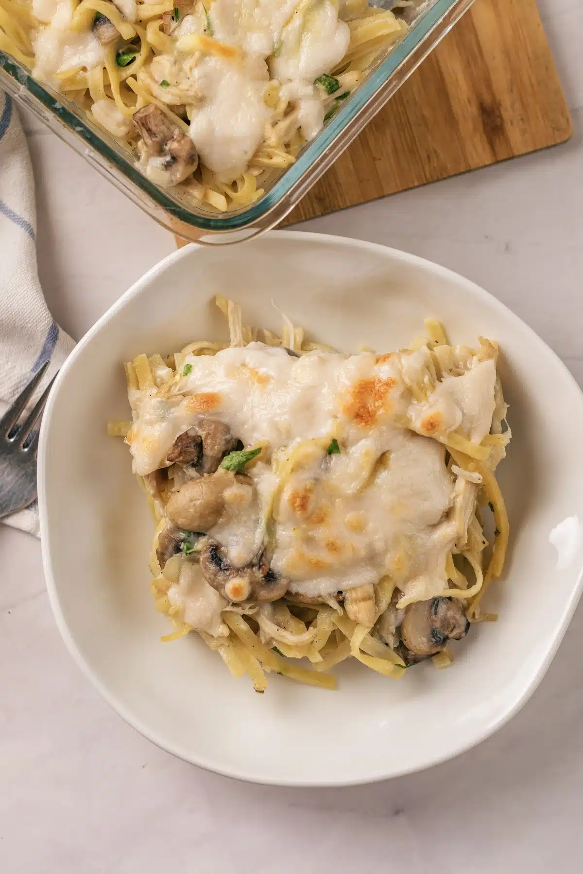 A square of chicken tetrazzini on a white plate with the glass dish and a fork behind it