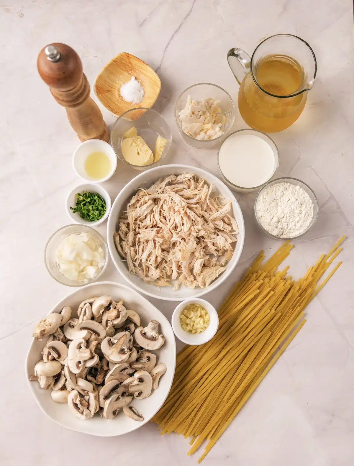 Shredded chicken, sliced mushrooms, linguine, broth, cream, butter, flour, onion, garlic, parsley and cheese in bowls on a marble counter