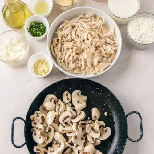 Sliced mushrooms cooking in the skillet with shredded chicken in a bowl behind