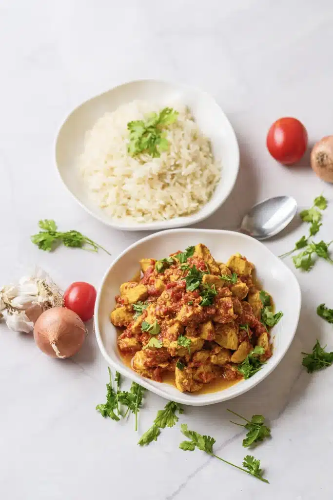 Chicken tikka masala on a white plate with a bowl of basmati rice, cilantro, garlic, an onion and a tomato, seen from above