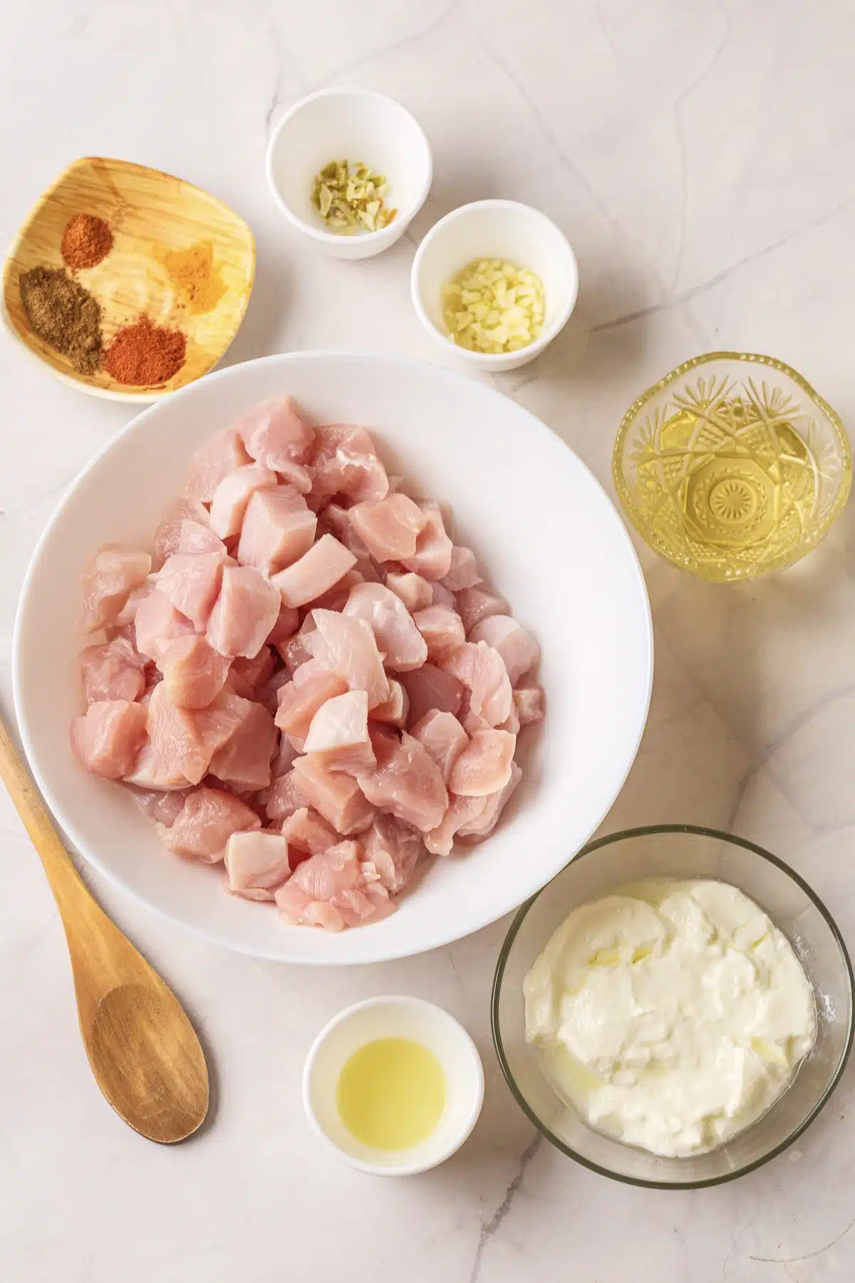 Cubed raw chicken in a white bowl with yogurt, ground spices, garlic, ginger, oil and lemon juice around it