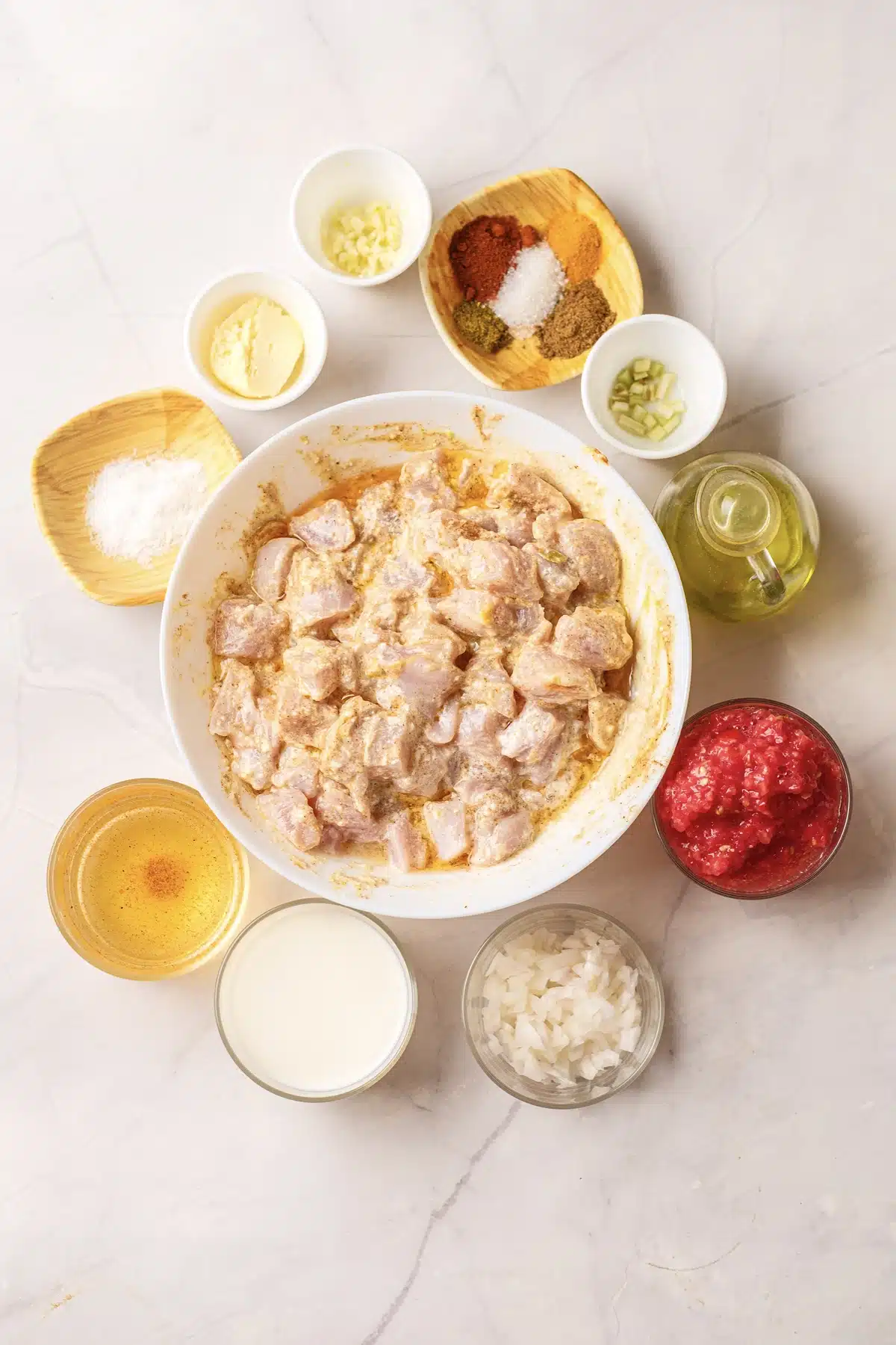 Marinated cubes in a bowl surrounded by crushed tomatoes, cream, chopped onion, ghee, oil and a dish of ground spices