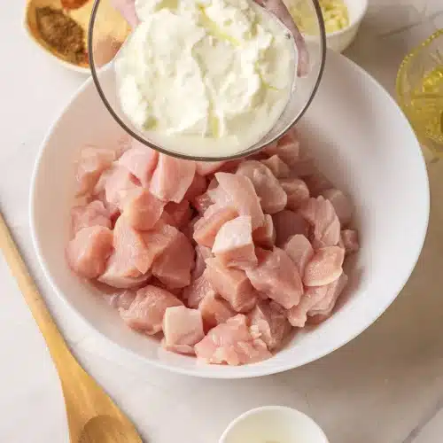 Thick yogurt tipped from a glass bowl onto cubed raw chicken in a white bowl