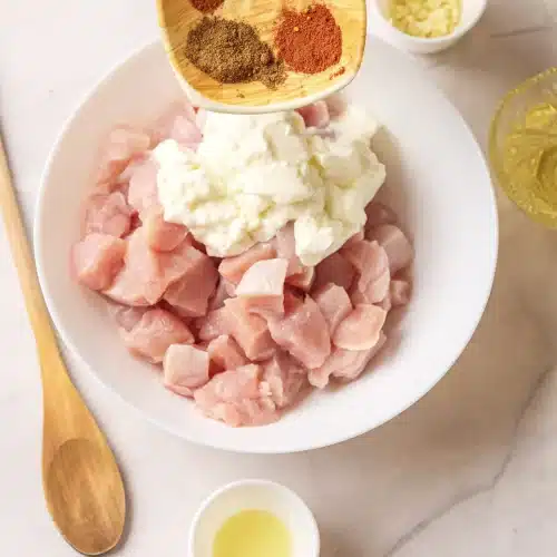 Ground spices poured from a small wooden dish onto the yogurt and chicken