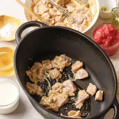 Marinated chicken pieces searing in a black cast-iron pot with tongs