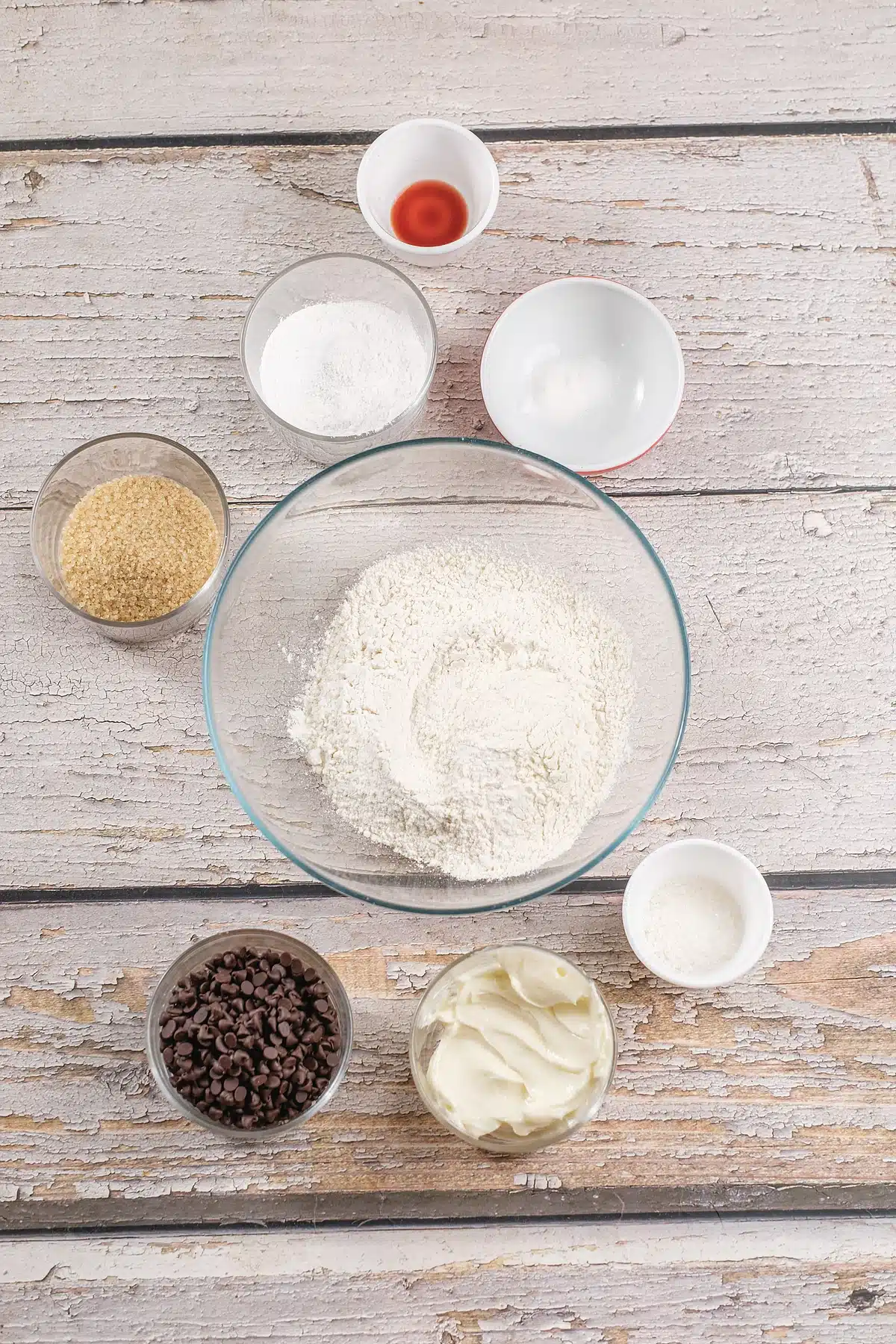 Flour, butter, brown sugar, eggs and dark morsels laid out in bowls on a white table