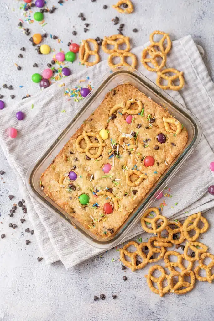 Christmas cookie bars in a glass dish with rainbow candy and mini pretzels on top, seen from directly above