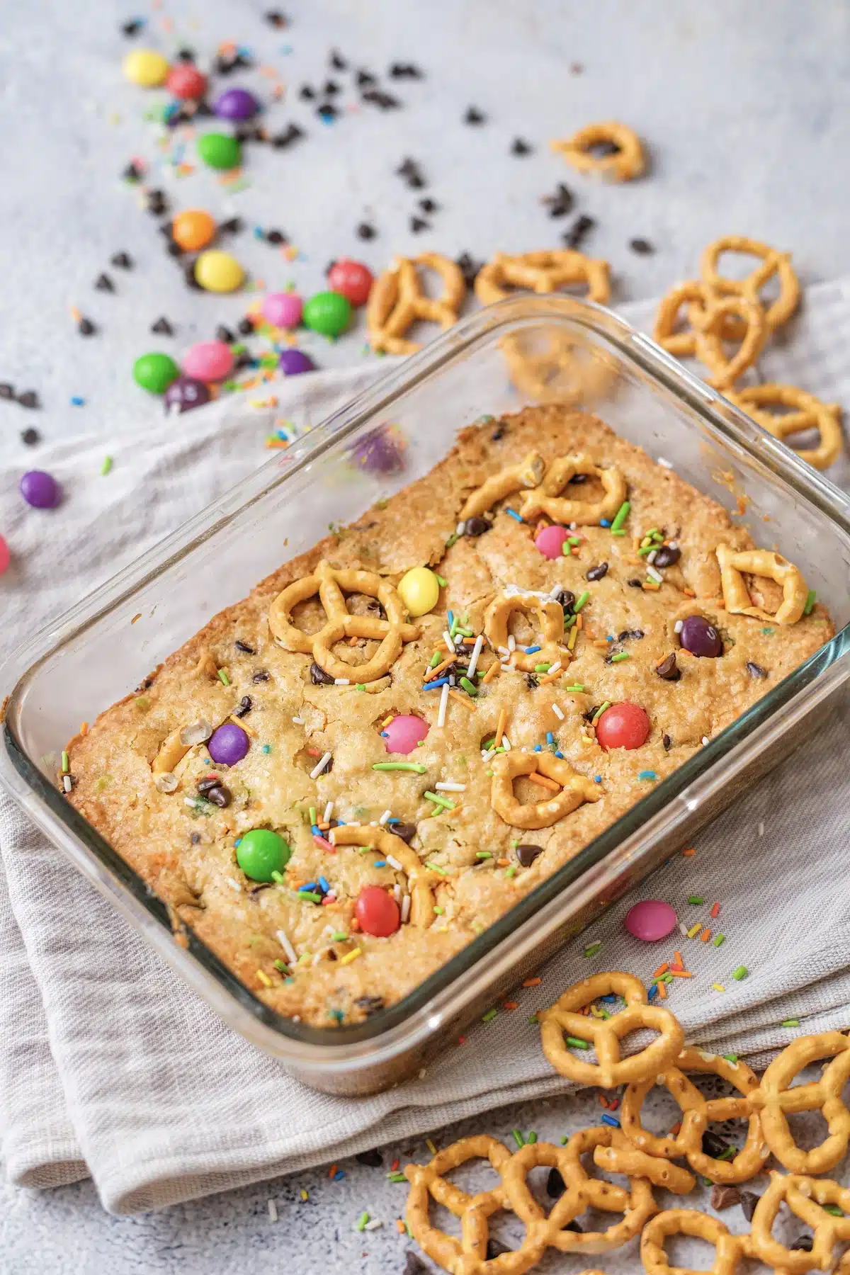Christmas cookie bars, rainbow version, baked whole in a glass dish with mini pretzels and candy scattered around it