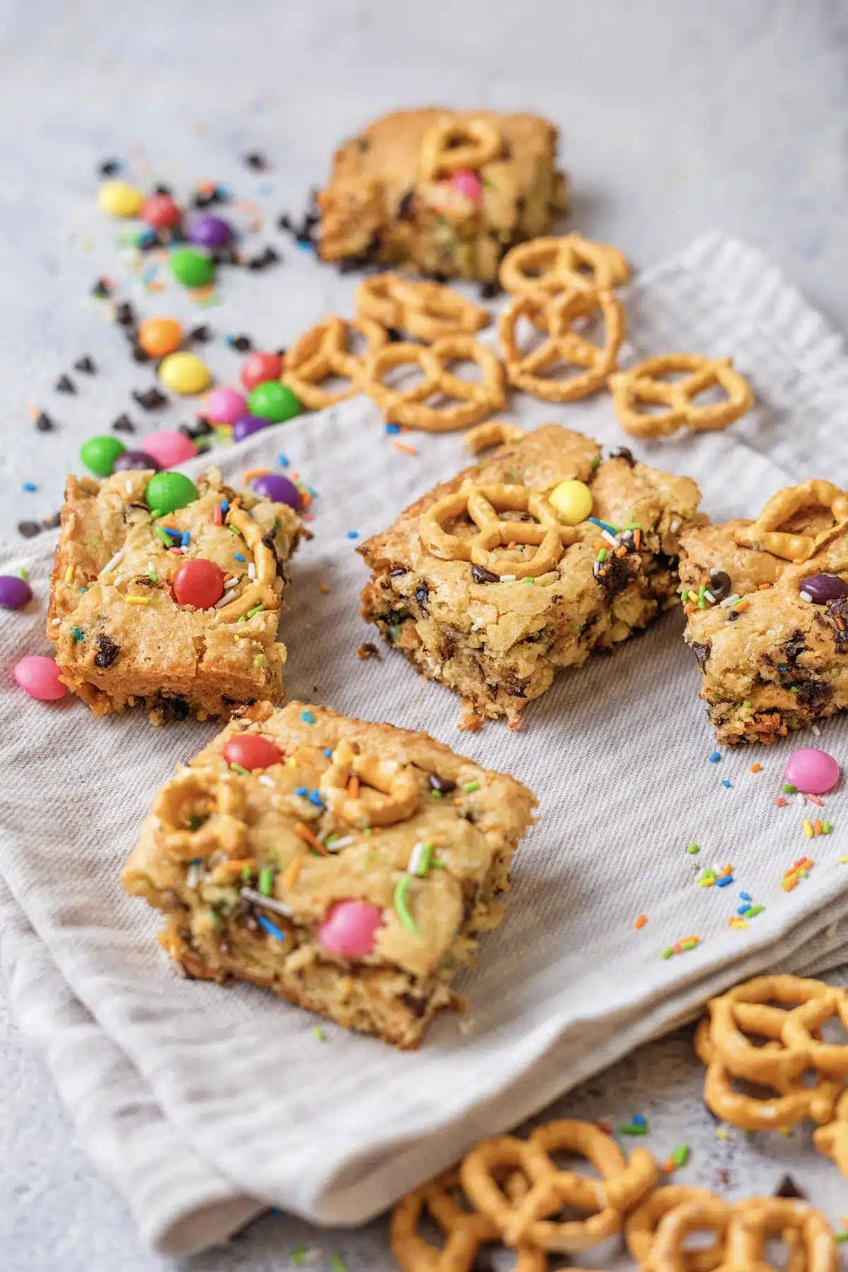 Christmas cookie bars in the rainbow version, stacked on a striped cloth with candy and pretzel twists beside them