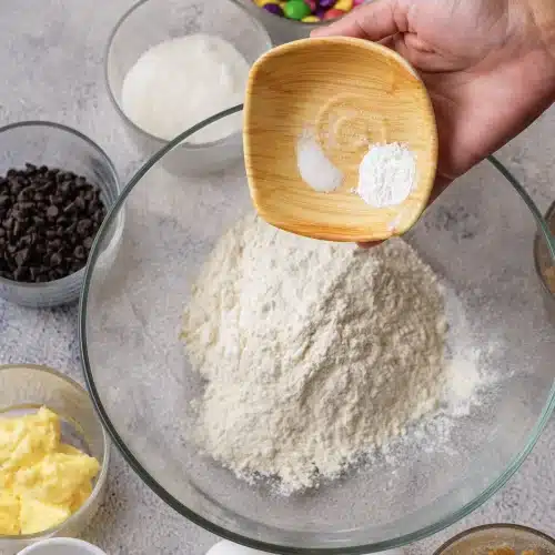 Baking powder and salt tipped from a small wooden bowl into a glass bowl of flour