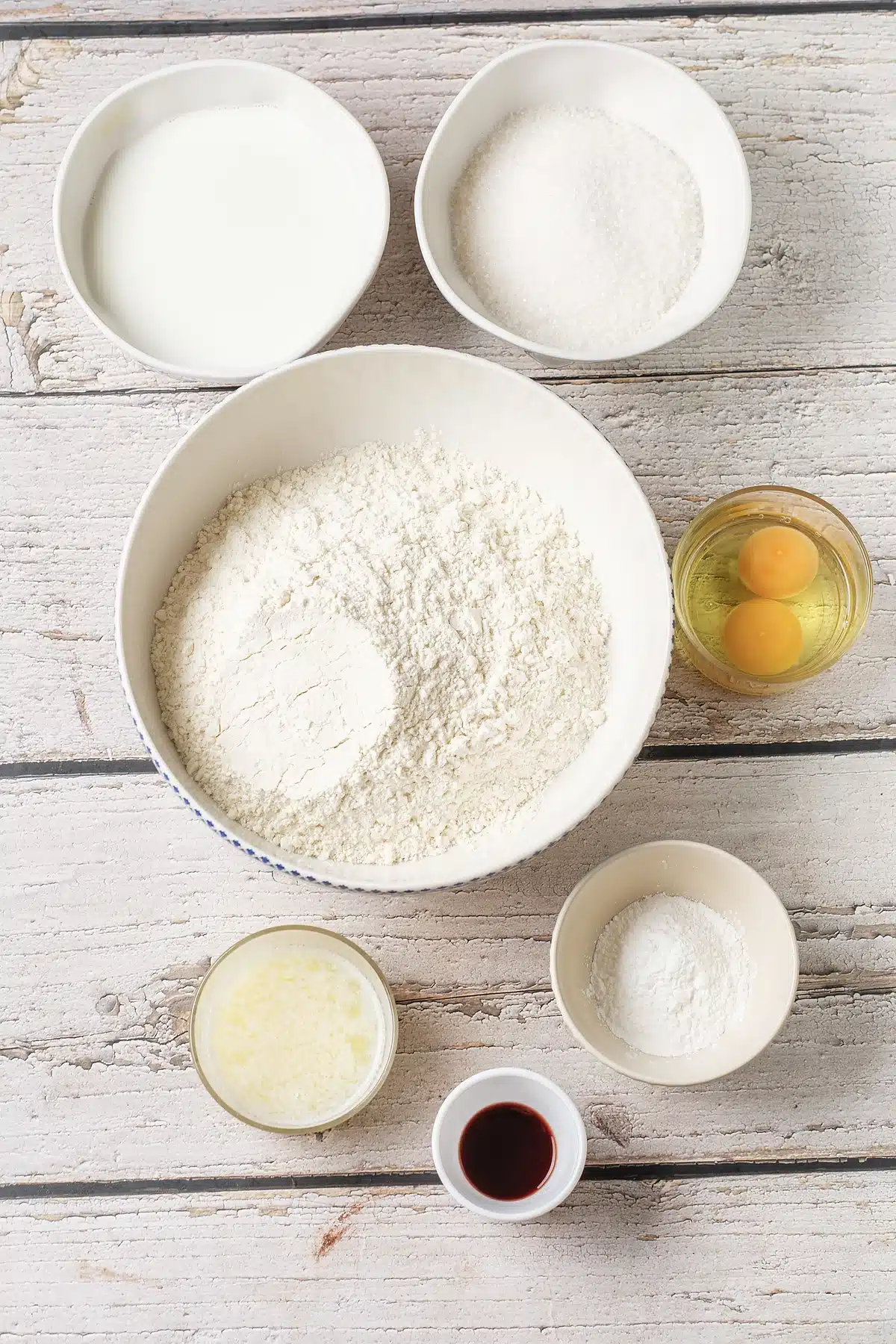 Flour, sugar, milk, eggs, butter and baking powder measured into white bowls