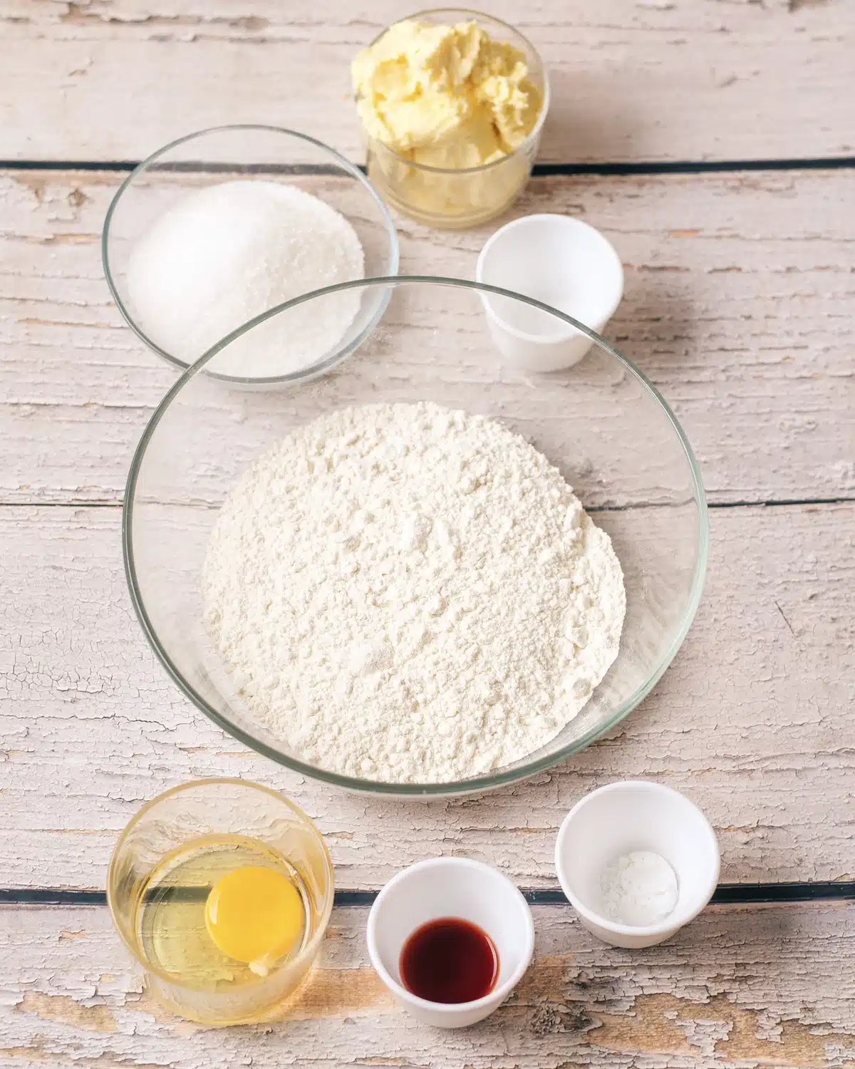 A large bowl of flour with softened butter, sugar, egg yolks, vanilla, a spoon of brown spice and salt around it