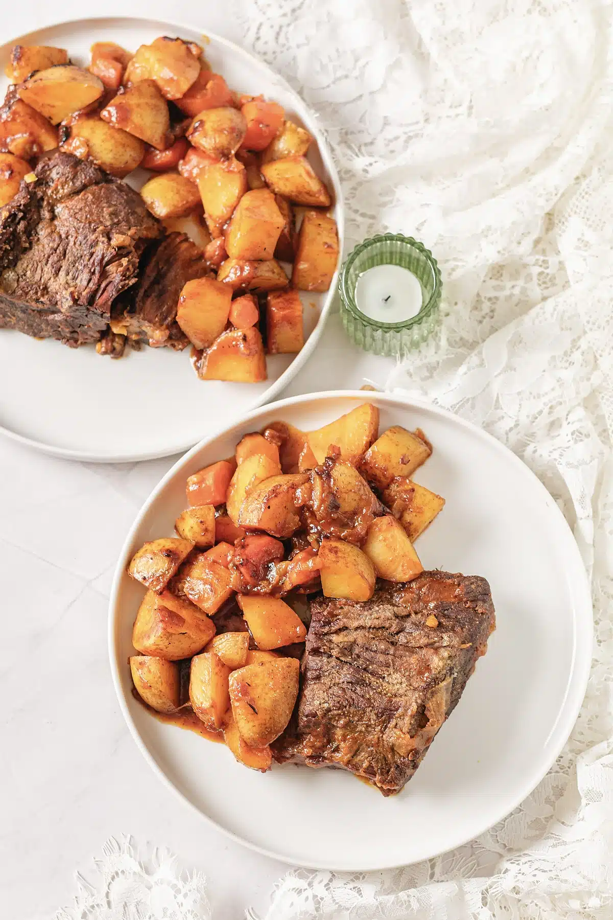 Overhead view of two white plates with beef and glazed vegetables beside a green candle