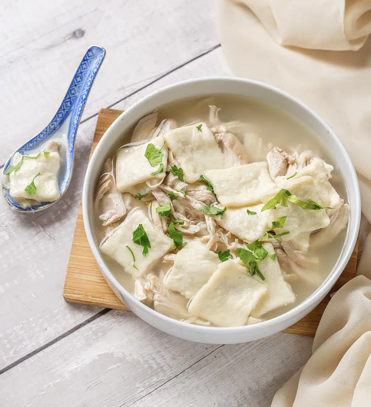A wide white bowl of thick broth with pale pasta-like squares and herbs