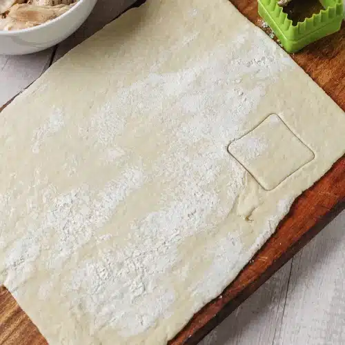 Rolled dough on a wooden board with one square cut out and a green cutter