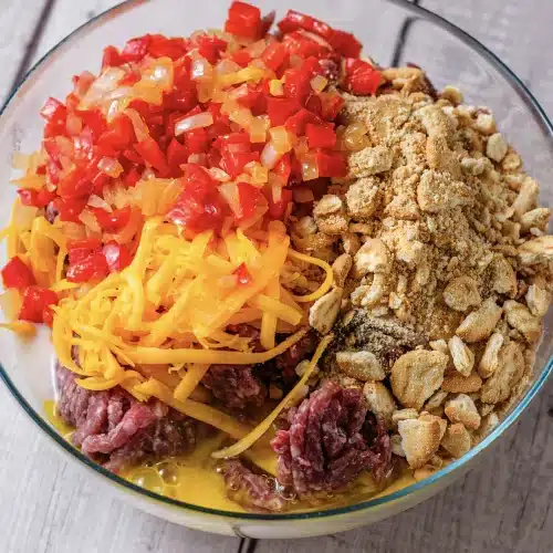 A glass bowl of raw ground beef heaped with shredded cheese, crumbs and diced pepper