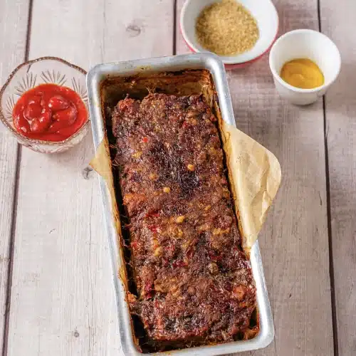 A baked brown loaf in its pan with small bowls of sugar and tomato sauce beside it
