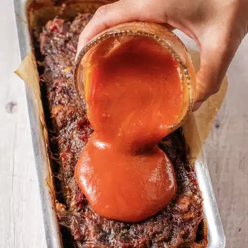 A hand tipping red glaze from a jar over the surface of a baked loaf