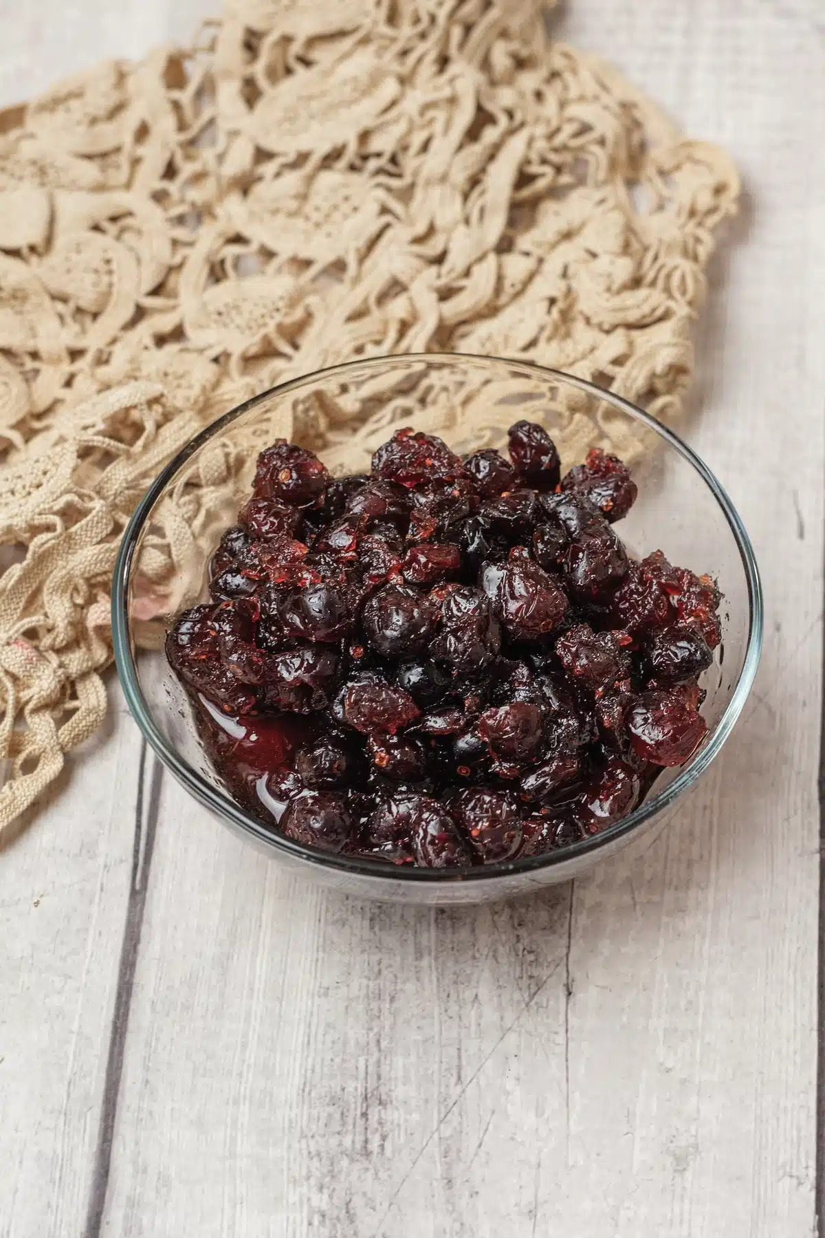 Whole dark berries in a thick red syrup heaped in a glass bowl on a lace cloth and whitewashed boards
