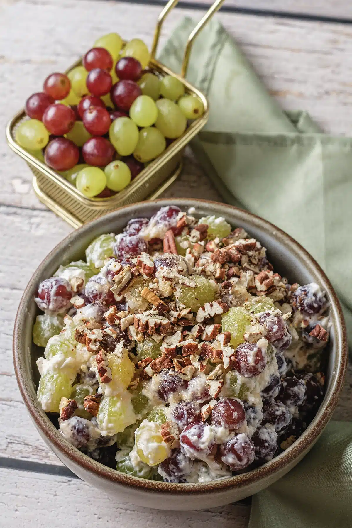 Grape salad in a stoneware bowl with a basket of red and green fruit behind it