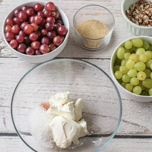 A glass bowl of softened cheese and sugar with bowls of fruit and pecans around it