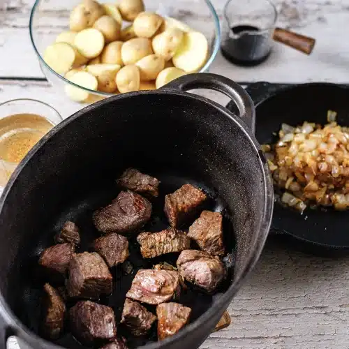Seared beef in a dark pot with halved potatoes and cooked onions waiting alongside