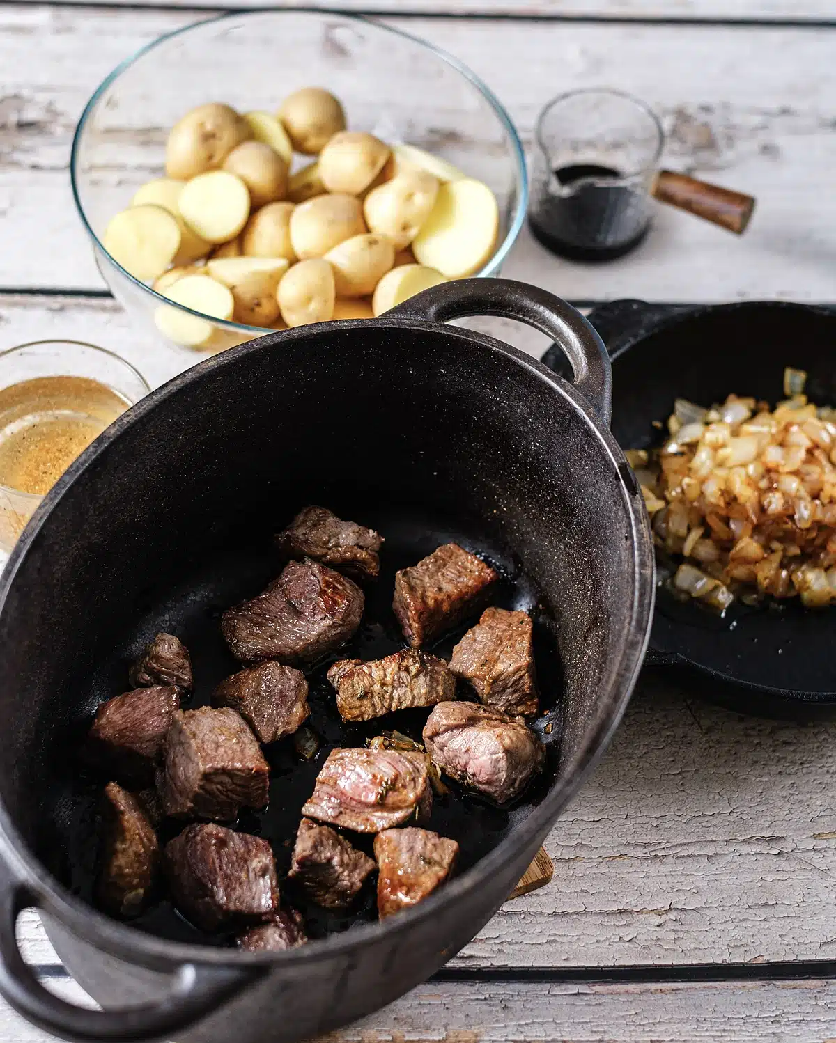 Seared beef in a dark pot with halved potatoes and cooked onions waiting alongside
