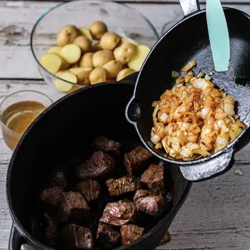 Cooked onions being tipped from a skillet into a dark pot of browned meat