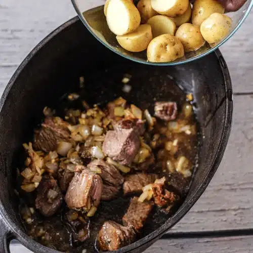 Hands adding halved baby potatoes to a dark pot of meat and onions