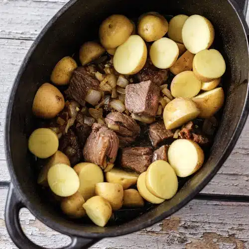 A dark oval pot loaded with raw halved potatoes over browned meat, ready to cook