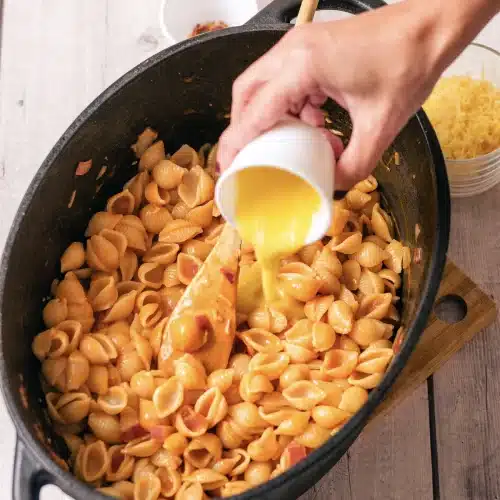 Starchy cooking water poured from a cup into the skillet of sauced shells